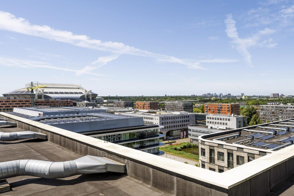 View over Elizabeth Cady Stantonplein with modern buildings and the Johan Cruijff ArenA in the background on a sunny day.